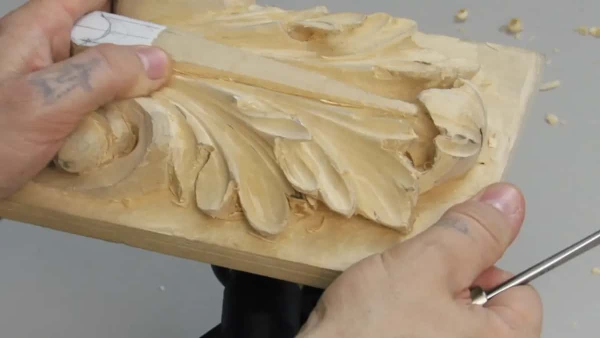 Holding up a carved Roman acanthus panel in plain wood, a thin steel tool in the other hand