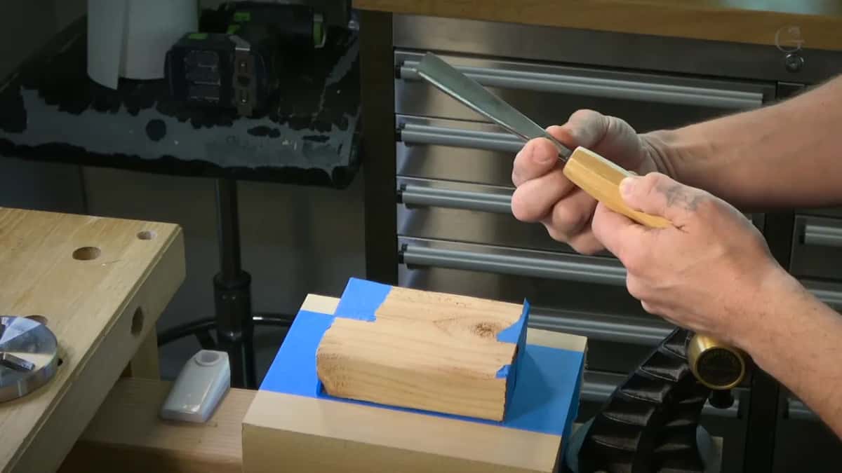 Hands turn a gouge with a faceted handle above a taped block