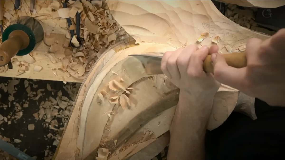 Two hands push a broad flat blade along the curved band under the scroll, facets covering the face above