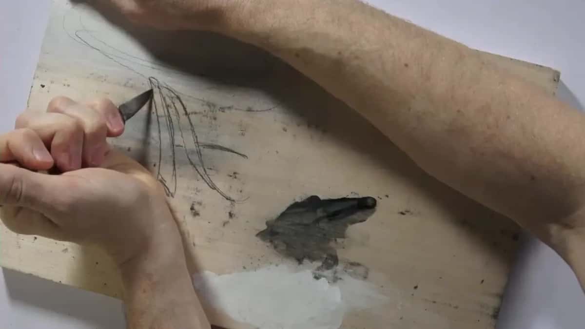 Knife tip sitting in the first pencil stroke while the other forearm rests across the top of the board