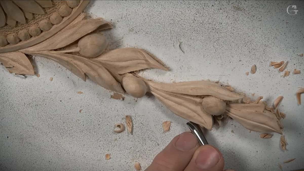 Beside a garland of carved leaves and berries on a white sheet, a small gouge rests between thumb and finger
