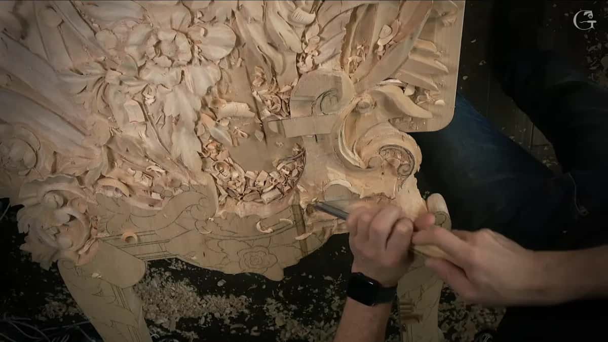 Both hands work a small tool close under a carved scroll, a drawn flower below