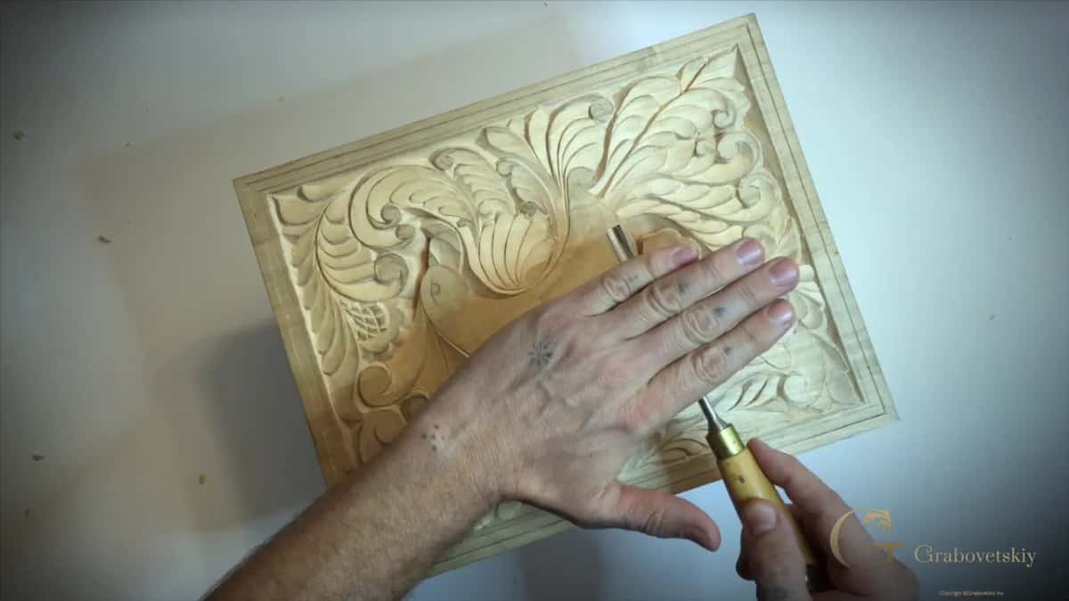 Flat over the blade, one hand rests on a carved leaf panel, the other holds the handle