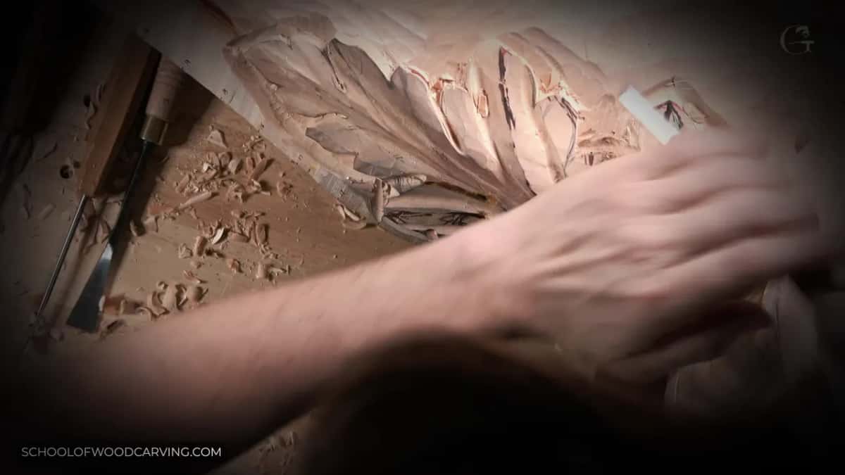 A hand rests across carved lobes while gouges wait on the bench, shavings everywhere