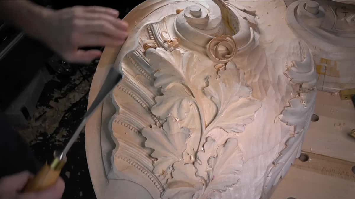 Two hands steer a long gouge past carved leaves, beads and a scroll