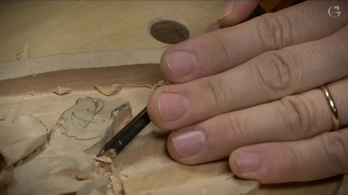 Seen low across the panel, fingers guide a gouge into the drawn bud while chips pile up