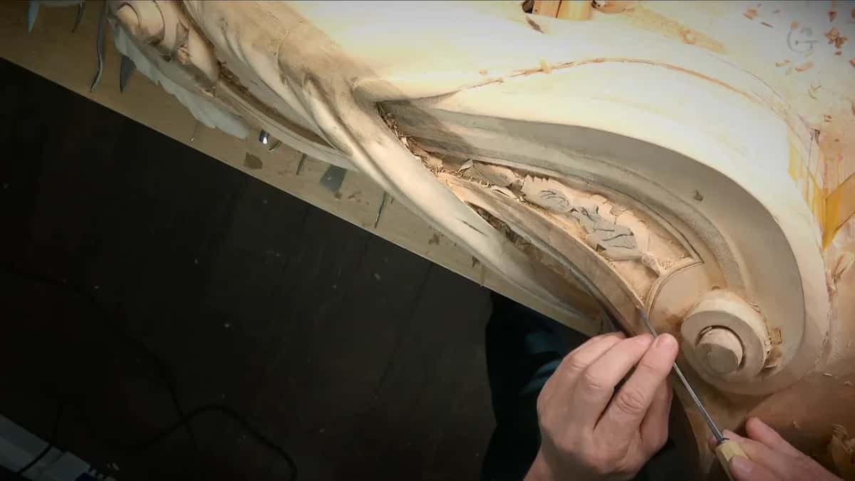 Both hands guide a slim tool along the inside curve of a carved scroll.